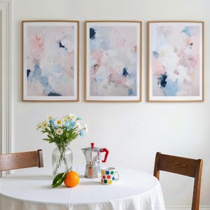 Puede incluir: Tres impresiones art&iacute;sticas abstractas enmarcadas con marcos de madera clara cuelgan sobre una mesa. El arte presenta suaves pinceladas rosas, azules y blancas. Un jarr&oacute;n de margaritas, una cafetera, una taza con lunares de colores y una naranja est&aacute;n sobre la mesa.