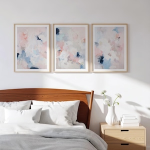 Puede incluir: Tres impresiones art&iacute;sticas abstractas enmarcadas. Cada impresi&oacute;n presenta un dise&ntilde;o suave y mezclado con tonos pastel rosa, azul y blanco. Los marcos son de color madera clara, y la obra de arte se exhibe sobre una cama.