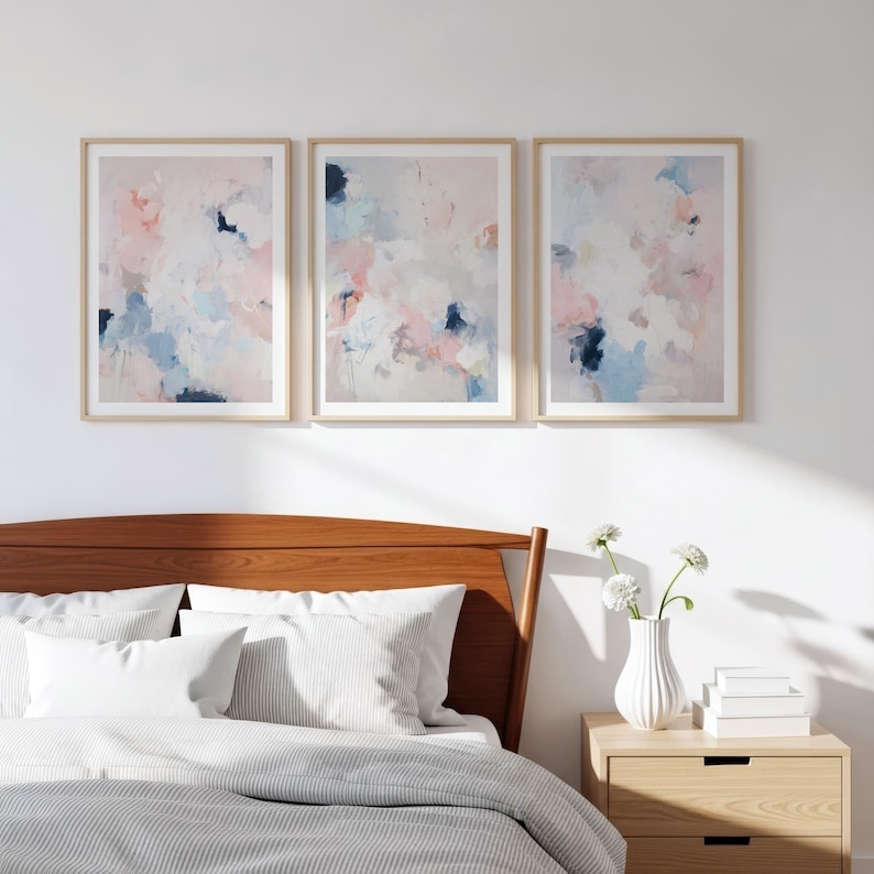 Puede incluir: Tres impresiones art&iacute;sticas abstractas enmarcadas. Cada impresi&oacute;n presenta un dise&ntilde;o suave y mezclado con tonos pastel rosa, azul y blanco. Los marcos son de color madera clara, y la obra de arte se exhibe sobre una cama.