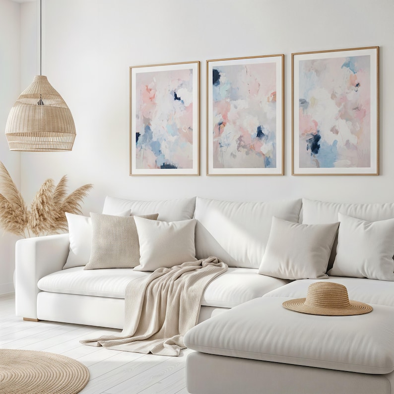 Puede incluir: Tres impresiones art&iacute;sticas abstractas enmarcadas con pinceladas rosas, azules y blancas cuelgan sobre un sof&aacute; blanco. La habitaci&oacute;n est&aacute; decorada con tonos neutros, incluyendo una l&aacute;mpara colgante tejida y una manta beige.