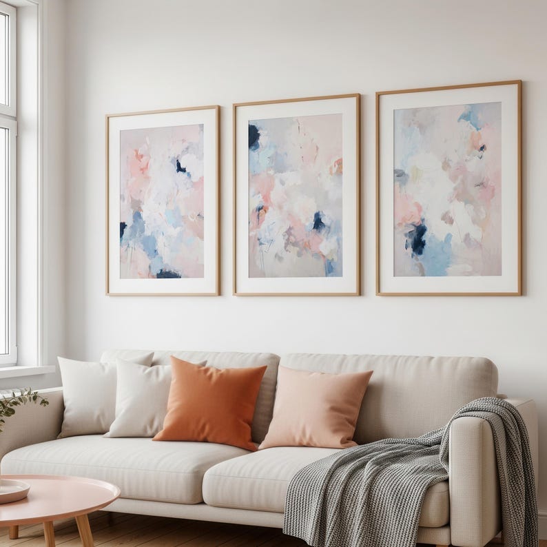 Puede incluir: Tres impresiones art&iacute;sticas abstractas enmarcadas. Cada impresi&oacute;n presenta una paleta de colores suaves de rosa, azul y blanco. Los marcos son de color madera clara, y la obra de arte cuelga sobre un sof&aacute; beige con cojines naranjas y melocot&oacute;n.