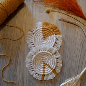 May include: Two round crocheted coasters in a white and mustard yellow colour scheme. The coasters have a fringe border and are stacked on top of each other.