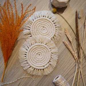 May include: Two white crocheted coasters with fringed edges. The coasters are round and have a woven pattern in the centre. The coasters are on a wooden surface with dried flowers and a spool of twine.