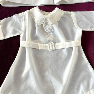 May include: A vintage white baby outfit, featuring a dress with a Peter Pan collar and a matching jacket. The dress has a decorative embroidered design on the bodice and a belt with a buckle. The outfit is made of a sheer, lightweight fabric.