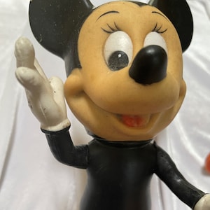 May include: A vintage Minnie Mouse figurine. The figure has a black body and ears, a tan face, and white gloves. The figurine is smiling and has a raised hand.