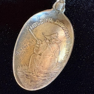 Puede incluir: Cuchara de plata con un diseño grabado. El cuenco de la cuchara presenta una imagen de una persona sosteniendo una caña de pescar, con las palabras "HOLDING DOWN A CLAIM" y "BLACKFOOT" grabadas alrededor de la imagen. El mango tiene un patrón decorativo.