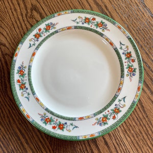 May include: White ceramic plate with a green border and floral design. The plate features a repeating pattern of orange, blue, and yellow flowers with green leaves. The plate is set against a wooden surface.