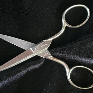 May include: Silver-toned scissors with curved handles and pointed blades, measuring approximately 4 inches. The blades are open, revealing the sharp edges. The scissors are set against a dark, textured background.