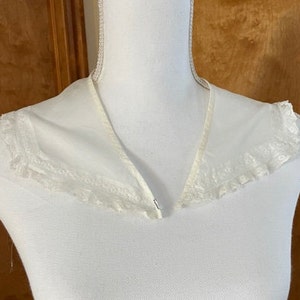 May include: A white, sheer collar with delicate lace trim. The collar is V-shaped and appears to be made of a lightweight fabric. The lace trim adds a vintage or antique aesthetic to the garment.