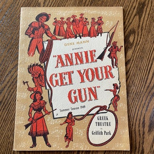 May include: Vintage "Annie Get Your Gun" playbill from the Greek Theatre in Griffith Park, Summer Season 1949. The cover features illustrations of cowboys, cowgirls, and Native American figures in red and black on a tan background.