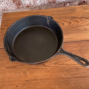 May include: A black cast iron skillet with a long handle and a rounded cooking surface. The skillet is sitting on a wooden surface. The handle has a hole for hanging. The skillet is a kitchenware item.