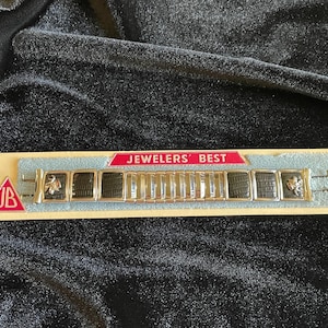 Può includere: Un braccialetto vintage con una fascia in metallo color argento e inserti rettangolari neri. Il braccialetto è esposto su una scheda azzurra con la scritta "JEWELERS' BEST" e un triangolo rosso con le lettere "JB".