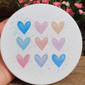 May include: A white ceramic coaster with a splatter paint design featuring nine pastel hearts in various shades of blue, pink, and purple.