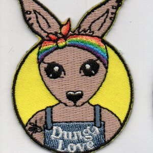 Peut inclure: Patch brodé représentant un kangourou de dessin animé portant une salopette avec le texte "Dunga Love" et un bandeau arc-en-ciel. Le kangourou est entouré d'un cercle jaune.