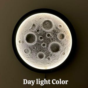 Op de afbeelding: Een witte maanvormige lamp met een zwart frame. Het licht is aan en verlicht het oppervlak van de maan, waardoor kraters en andere details zichtbaar worden. De tekst "Day light Color" staat onder de maan.