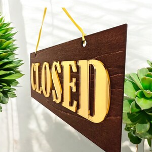 Op de afbeelding: Een bruin houten bord met een gouden "GESLOTEN"-bord dat aan een geel touwtje hangt. Het bord heeft een verweerde houten uitstraling.