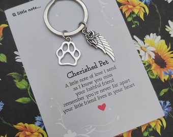 Puede incluir: Un llavero plateado con un dije de huella de pata, una rosa y un dije de ala. El llavero está unido a una tarjeta con el texto "Cherished Pet" y un mensaje sobre recordar a un ser querido.