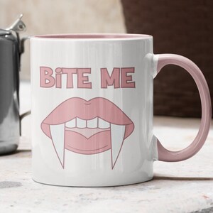 Peut inclure: Une tasse en c&eacute;ramique blanche avec un bord et une anse roses. La tasse porte l'inscription "Bite Me" en lettres roses et une image de dessin anim&eacute; rose d'une bouche de vampire avec des crocs.