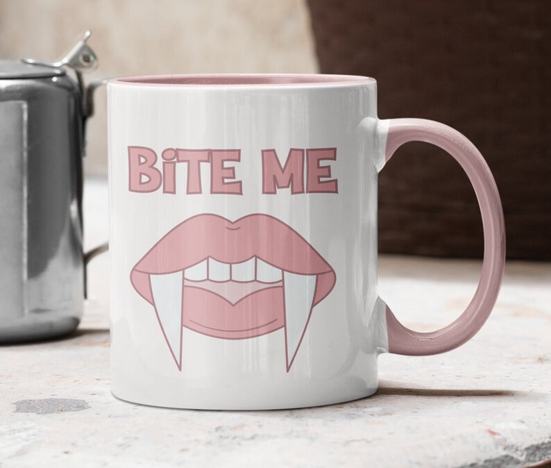 Peut inclure: Une tasse en c&eacute;ramique blanche avec un bord et une anse roses. La tasse porte l'inscription "Bite Me" en lettres roses et une image de dessin anim&eacute; rose d'une bouche de vampire avec des crocs.