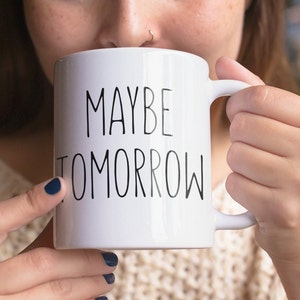 May include: White ceramic coffee mug with black text that reads "MAYBE TOMORROW".