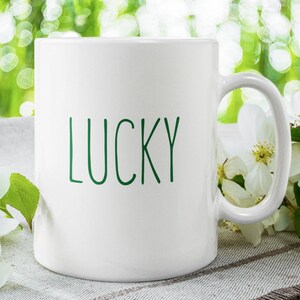 May include: White ceramic mug with a green inscription that reads "LUCKY". The mug is sitting on a white and brown fabric surface with white flowers.
