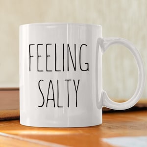 May include: White ceramic mug with the words "FEELING SALTY" printed in black, sans-serif font. The mug has a curved handle and is sitting on a wooden surface. The mug is a standard size for coffee or tea.