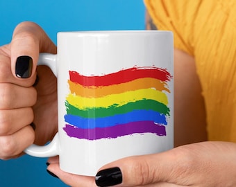 May include: White ceramic mug with a rainbow flag design painted in a brushstroke style. The flag is red, orange, yellow, green, blue, and purple.