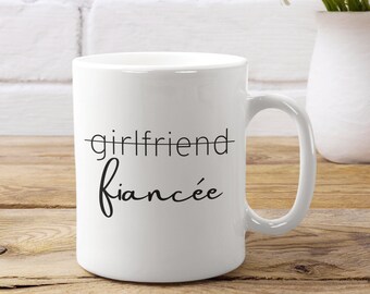 May include: White ceramic coffee mug with black text that reads "girlfriend" with a line through it and "fiancée" written below it.