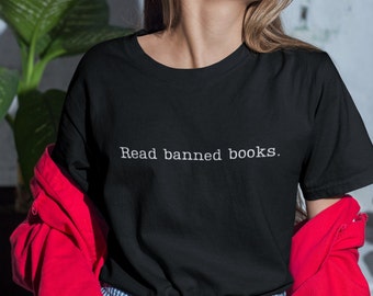 May include: Black t-shirt with the text "Read banned books." in a white, simple font. The shirt is a classic crew neck style. The person in the image is wearing a red jacket.
