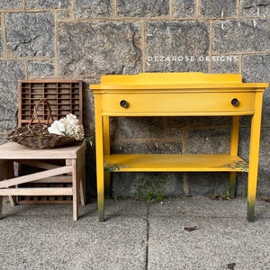 May include: A yellow painted side table with a single drawer and a lower shelf. The table has a distressed finish and is decorated with a floral pattern. The table is sitting in front of a stone wall.