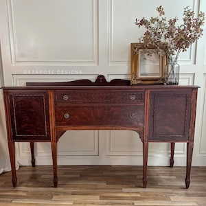 May include: A vintage mahogany wood sideboard with two drawers and a cabinet door. The sideboard has intricate carvings and a rich, dark finish. The text "DEZAROSE DESIGNS" is visible on the top of the sideboard.
