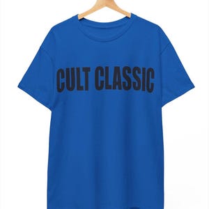 以下が含まれることがあります： 胸元に太字の黒い大文字で「CULT CLASSIC」というテキストがプリントされた、鮮やかなブルーの半袖Tシャツ。木製のハンガーに掛けられており、クルーネックと標準的なストレートの裾が特徴です。
