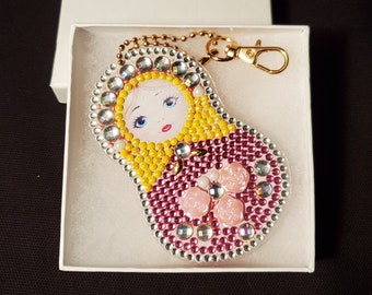 May include: A Matryoshka doll-shaped keychain with a gold-colored clasp and chain. The doll features a painted face, yellow hair, and a pink dress, embellished with clear and pink rhinestones. It is presented in a white gift box.
