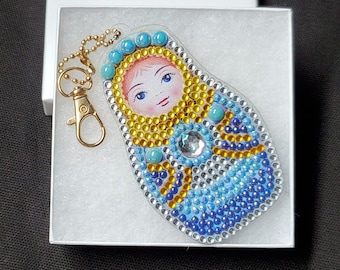 May include: A Russian nesting doll keychain with a gold-colored chain and clasp. The doll is decorated with blue, gold, and clear rhinestones, and features a painted face. The keychain is presented in a white gift box.