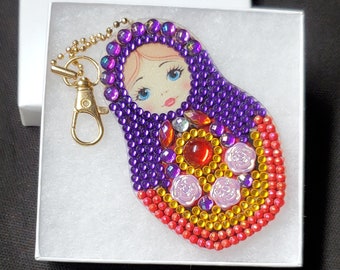 May include: A purple, red, and yellow beaded keychain shaped like a Russian nesting doll. The doll has a painted face with blue eyes and a gold chain.