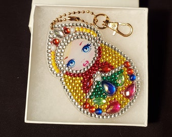 May include: A Matryoshka doll keychain with a gold-colored chain and clasp. The doll is decorated with colorful rhinestones in red, yellow, green, blue, and pink. The doll's face has blue eyes, a red mouth, and blonde hair. The keychain is in a white box.