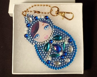 May include: A blue and white Matryoshka doll keychain with a gold-colored chain and clasp. The doll is decorated with blue and clear rhinestones, with a painted face and a white box.