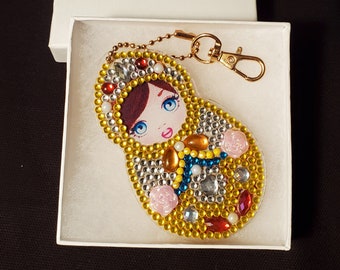 May include: A gold-colored keychain with a round, clear charm featuring a colorful, cartoon-style Russian nesting doll. The doll has a red, white, and blue patterned dress and a yellow background.