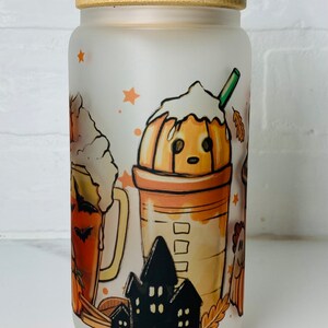 May include: A frosted glass jar with a bamboo lid features a Halloween-themed design. The design includes a pumpkin spice latte, a pumpkin beer, and a haunted house. The jar is perfect for storing treats or other small items.