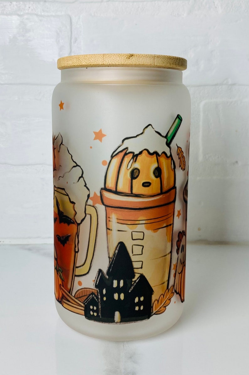 May include: A frosted glass jar with a bamboo lid features a Halloween-themed design. The design includes a pumpkin spice latte, a pumpkin beer, and a haunted house. The jar is perfect for storing treats or other small items.