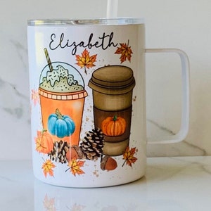 May include: White stainless steel travel mug with a handle. The mug features a fall-themed design with a pumpkin spice latte, a pumpkin, and autumn leaves. The mug is personalised with the name "Elizabeth".