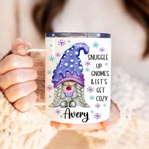 May include: White ceramic mug with a purple and white gnome holding a cupcake. The mug has snowflakes and the text "Snuggle Up Gnomes & Let's Get Cozy Avery" printed on it.