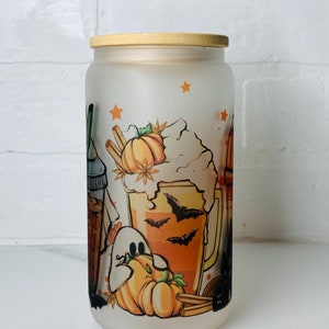 May include: A frosted glass jar with a wooden lid features a Halloween-themed design. The design includes a pumpkin, a ghost, a mug of orange liquid with bats, and cinnamon sticks.