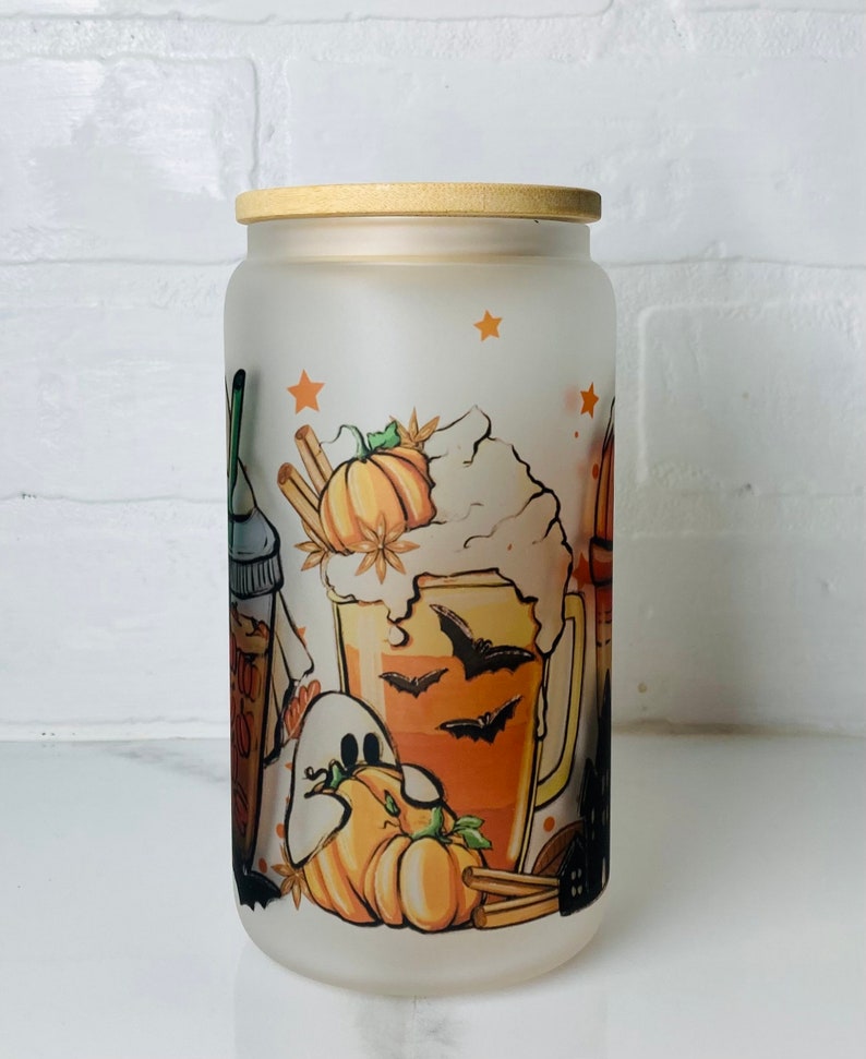 May include: A frosted glass jar with a wooden lid features a Halloween-themed design. The design includes a pumpkin, a ghost, a mug of orange liquid with bats, and cinnamon sticks.