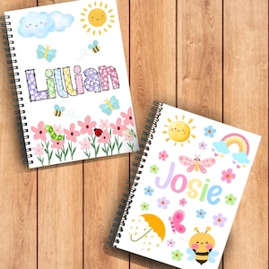 May include: Two spiral-bound notebooks with white covers. The first notebook features the name Lillian in patterned letters, a smiling sun, clouds, butterflies, bees, and pink flowers. The second notebook displays the name Josie, a smiling sun, a rainbow, butterflies, flowers, an umbrella, and a bee.
