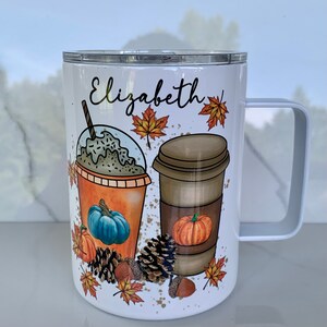 May include: White stainless steel tumbler with a white lid and a black handle. The tumbler features a fall-themed design with two coffee cups, pumpkins, and autumn leaves. The text "Elizabeth" is printed on the tumbler.