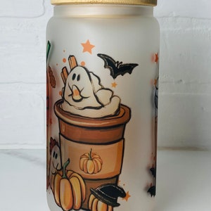 May include: A frosted glass jar with a bamboo lid features a whimsical Halloween-themed design. The jar has a white background with illustrations of a coffee cup with whipped cream topped with a ghost, pumpkins, bats, and stars.