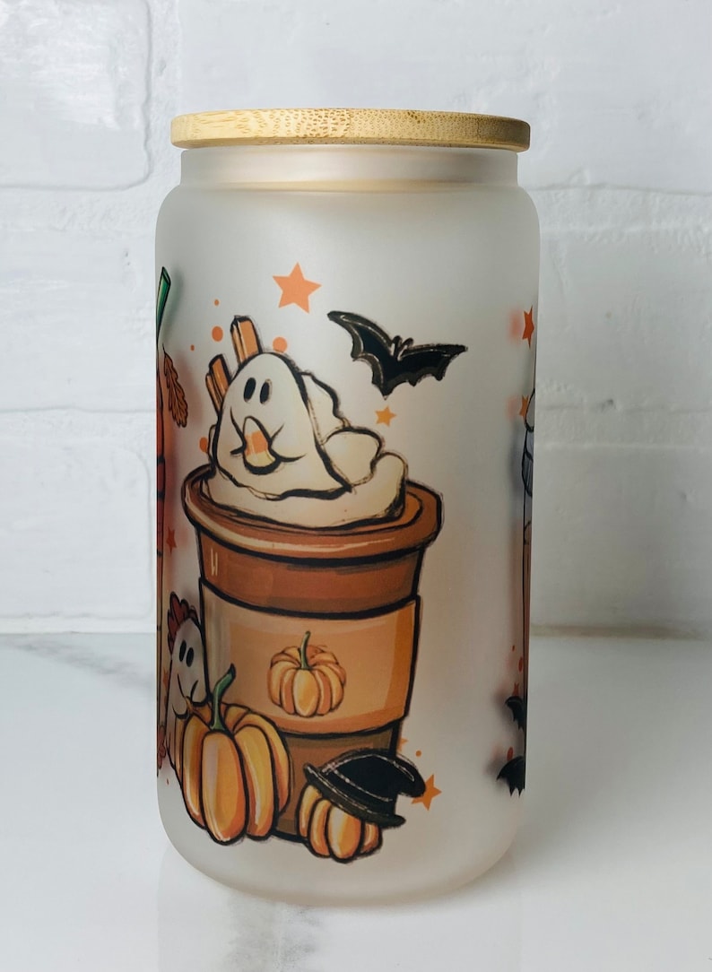 May include: A frosted glass jar with a bamboo lid features a whimsical Halloween-themed design. The jar has a white background with illustrations of a coffee cup with whipped cream topped with a ghost, pumpkins, bats, and stars.