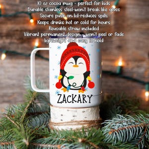 Może przedstawiać: Biały kubek do kakao o pojemności 295 ml z motywem pingwina w czerwonej czapce i trzymającego lampki choinkowe. Na kubku widnieje imię "ZACKARY". Wykonany jest ze stali nierdzewnej i zawiera słomkę wielokrotnego użytku.
