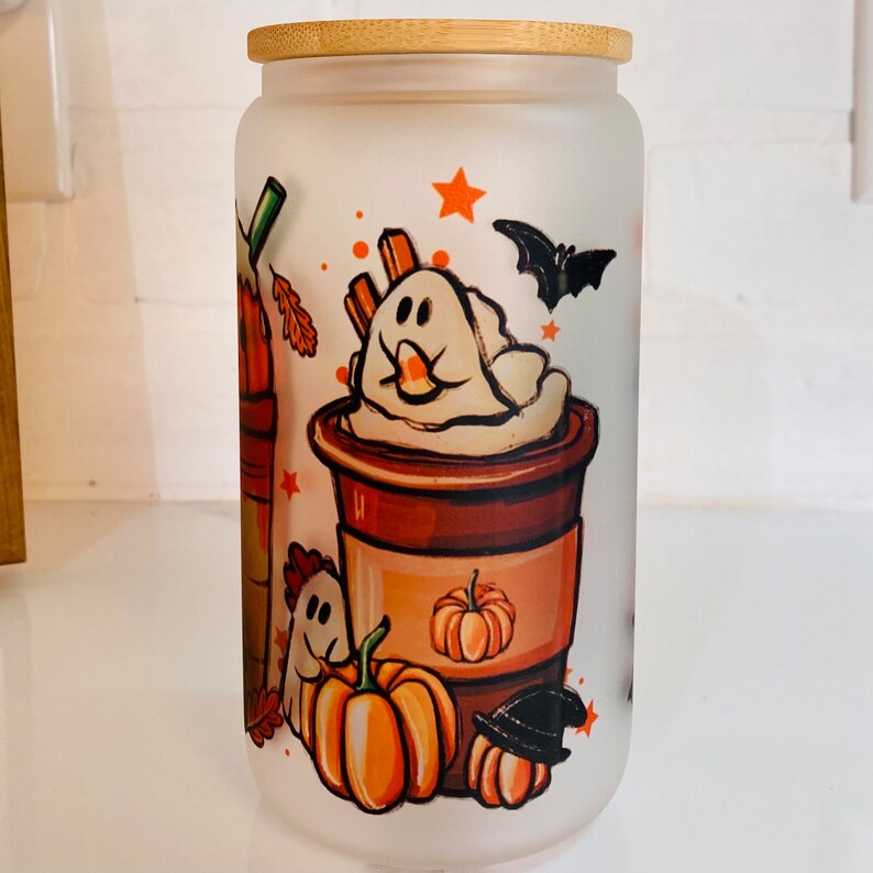May include: A frosted glass jar with a bamboo lid features a Halloween-themed design. The design includes a ghost, pumpkins, and a bat. The jar is perfect for storing treats or other small items.